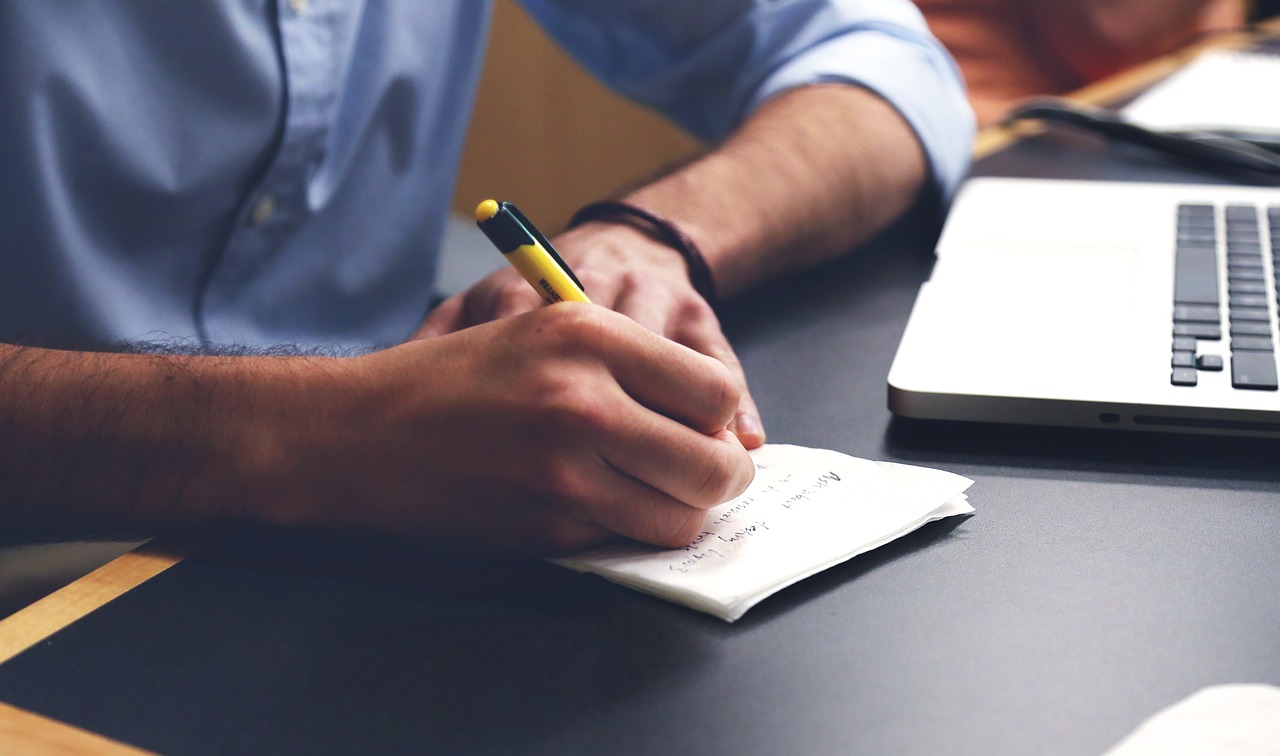 Uma mão masculina escrevendo em um papel em cima de uma mesa e do lado um notebook.