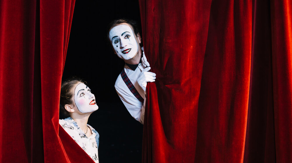Um casal de artista sorrindo atrás de uma cortina de um palco de teatro