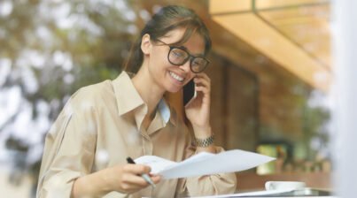 Mulher de negócios feliz examinando documento e falando no celular enquanto está sentada à mesa num café.
