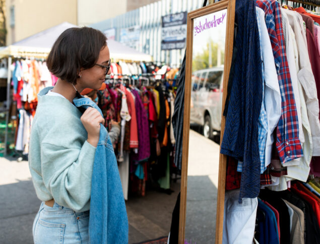 Uma mulher vendo uma camiseta na frente de um espelho de um comércio na rua.