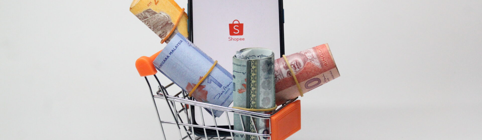 Miniatura de um carrinho de supermercado com notas de dinheiro e um celular mostrando o logo da Shopee