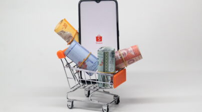 Miniatura de um carrinho de supermercado com notas de dinheiro e um celular mostrando o logo da Shopee